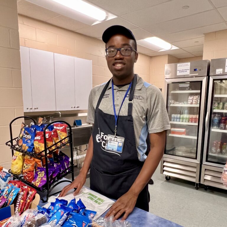 Focus Cafe team member standing behind a counter with snacks and refrigerated drinks